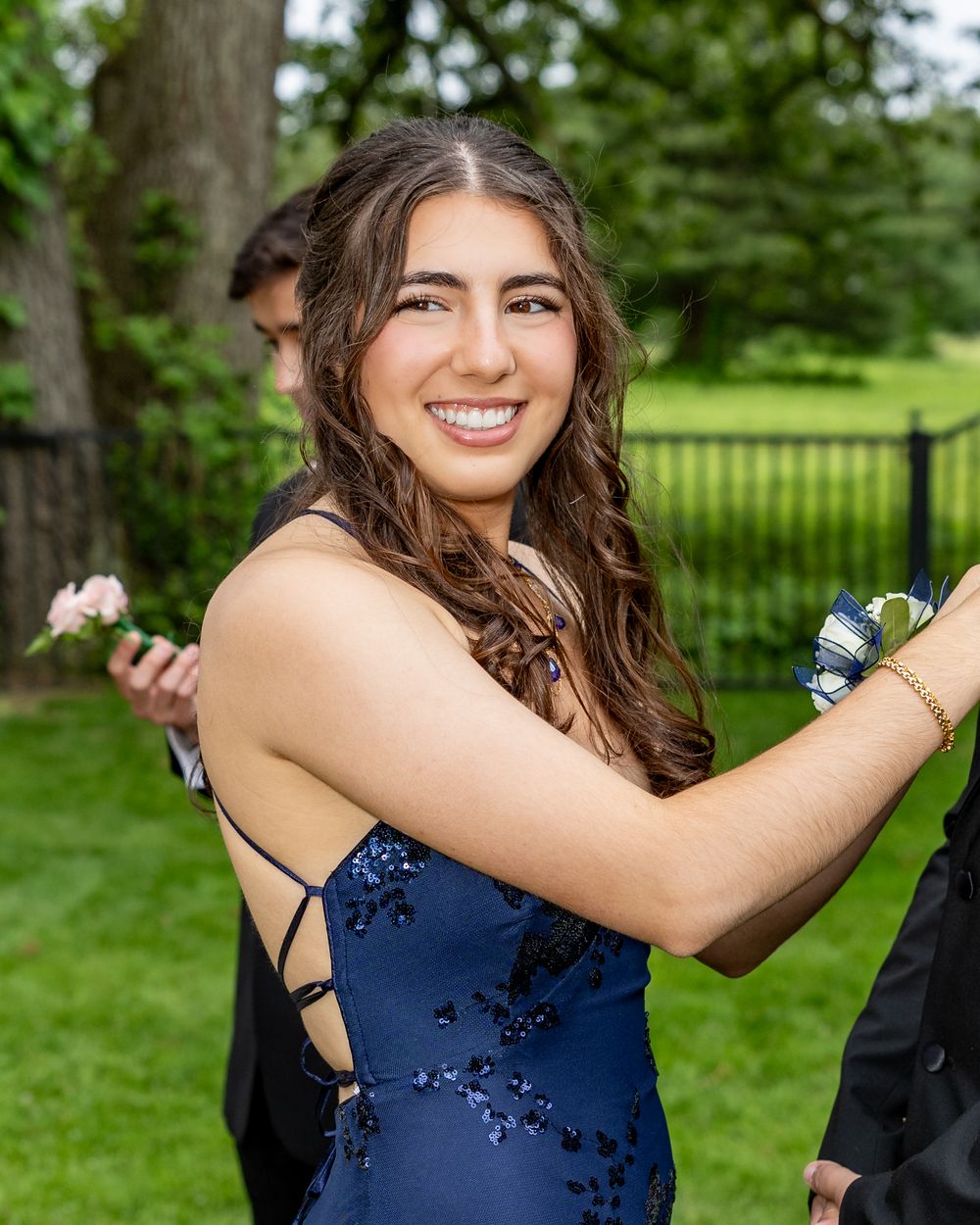 A smiling young woman in a navy blue sequin prom dress with a floral wrist corsage poses outdoors at pre prom party photos in Concord, MA.