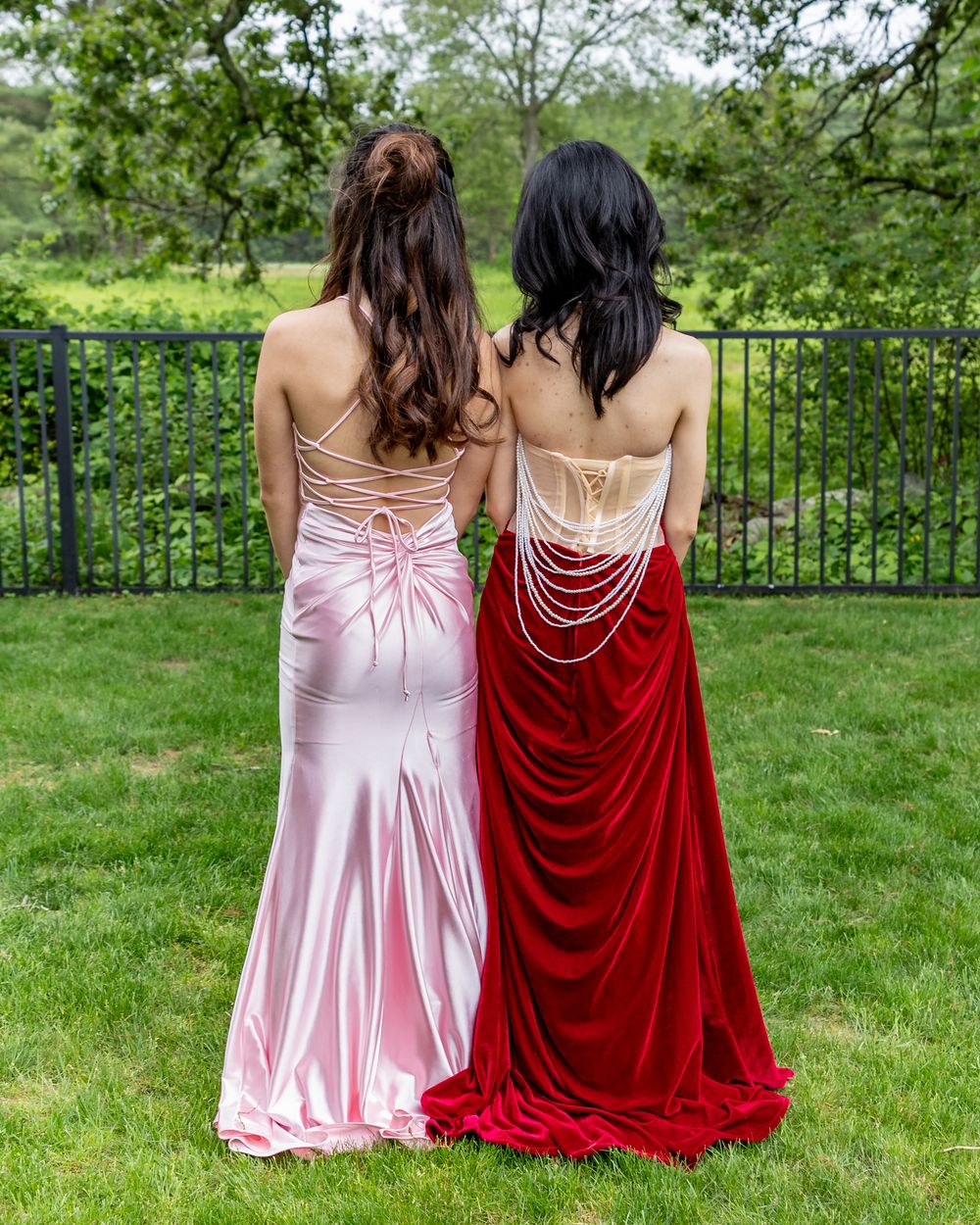 Two women in elegant backless prom gowns, one pink satin and one red velvet with pearl draping, posing outdoors at pre prom photos in Weston
