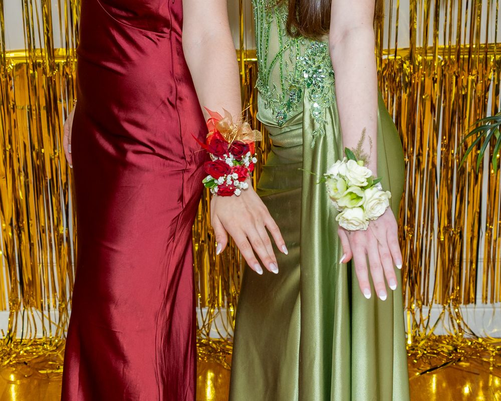 Two girls in elegant red and green satin prom gowns displaying floral wrist corsages against a gold backdrop at pre prom party .