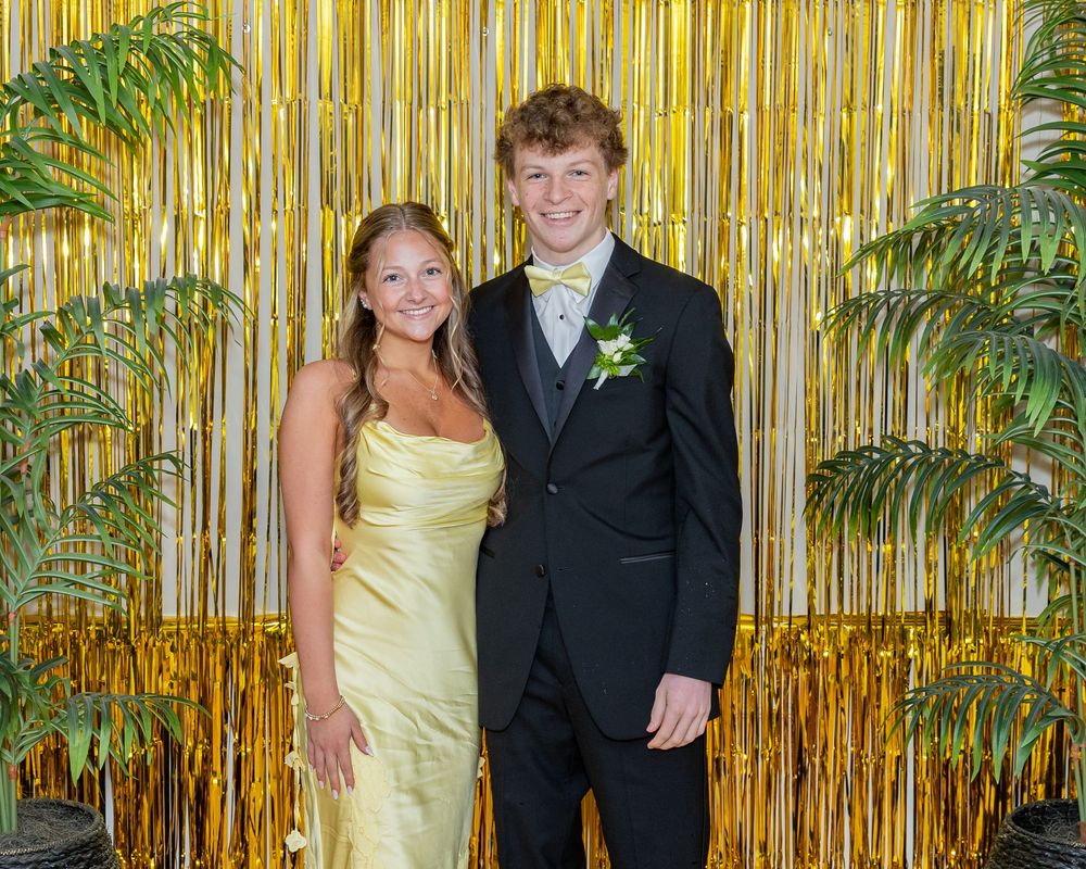Teenage couple posing at prom in front of gold tinsel backdrop, wearing formal attire with corsage and boutonniere at pre prom party.