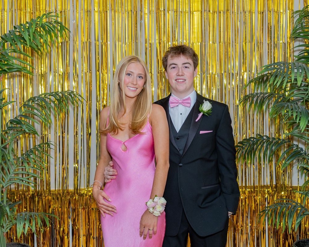A young couple poses in prom attire, she in pink gown, he in black tuxedo, before a gold tinsel backdrop at pre prom photos outside Boston