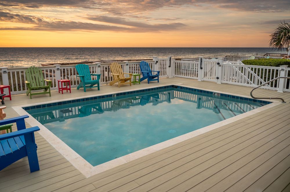 Colorful Adirondack chairs line a wooden deck overlooking a swimming pool at sunset with palm trees in the background.