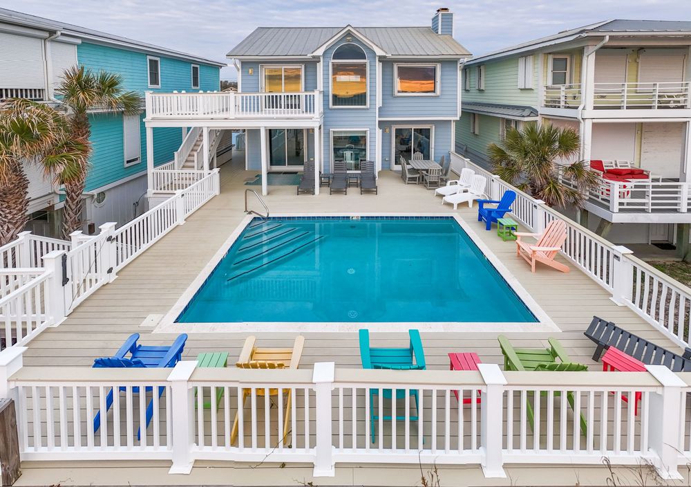 Coastal beach house with gray exterior, white deck railings and large swimming pool featuring colorful lounge chairs.