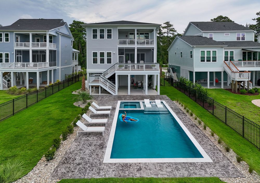 A panoramic view of a modern backyard featuring a rectangular swimming pool surrounded by green lawn and white houses.