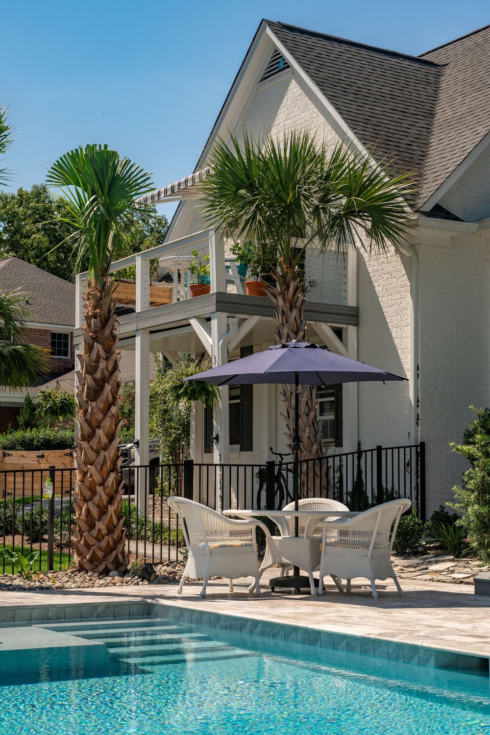 Modern backyard swimming pool with palm trees, white patio furniture, and a purple umbrella next to a contemporary house.