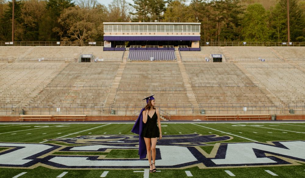 A graduate in cap and gown stands at the center of an empty football stadium with bleachers and press box visible.