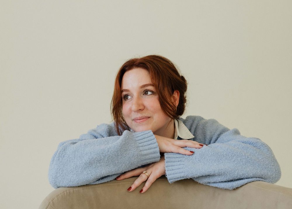Someone wearing a cozy gray sweater rests their chin on crossed arms while gazing upward with a gentle smile.