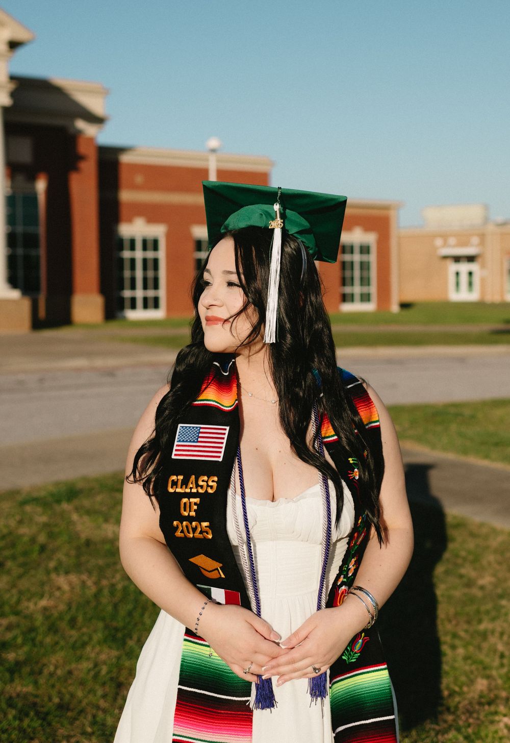 A graduate in cap and gown with colorful stoles poses outside a brick school building on graduation day.