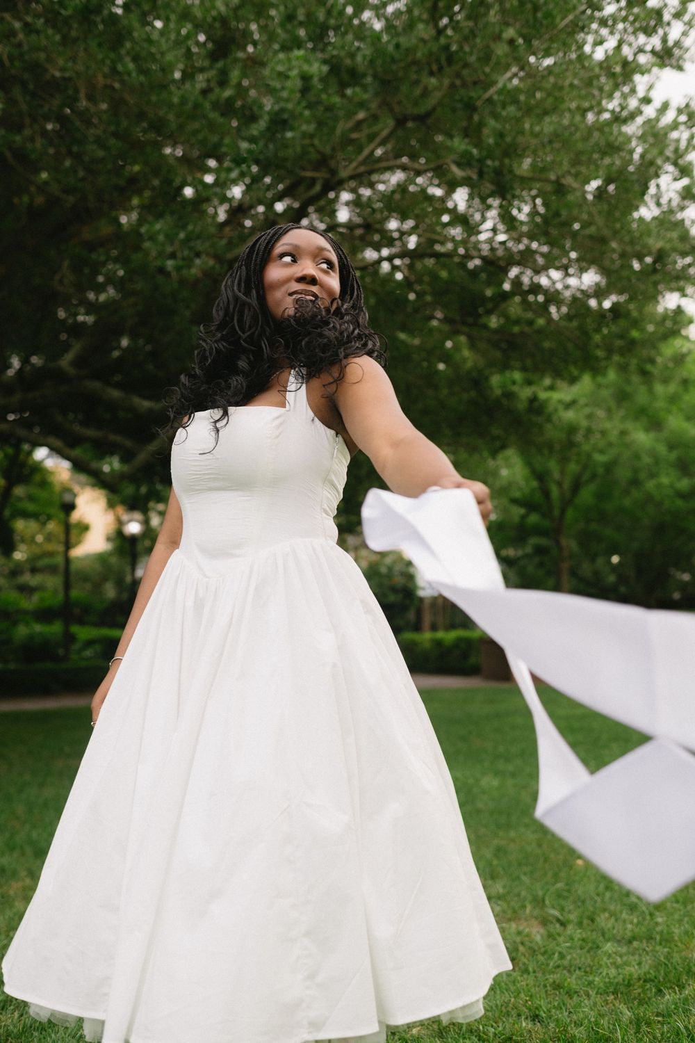 A person in a flowing white dress twirls joyfully on green grass in a garden setting.