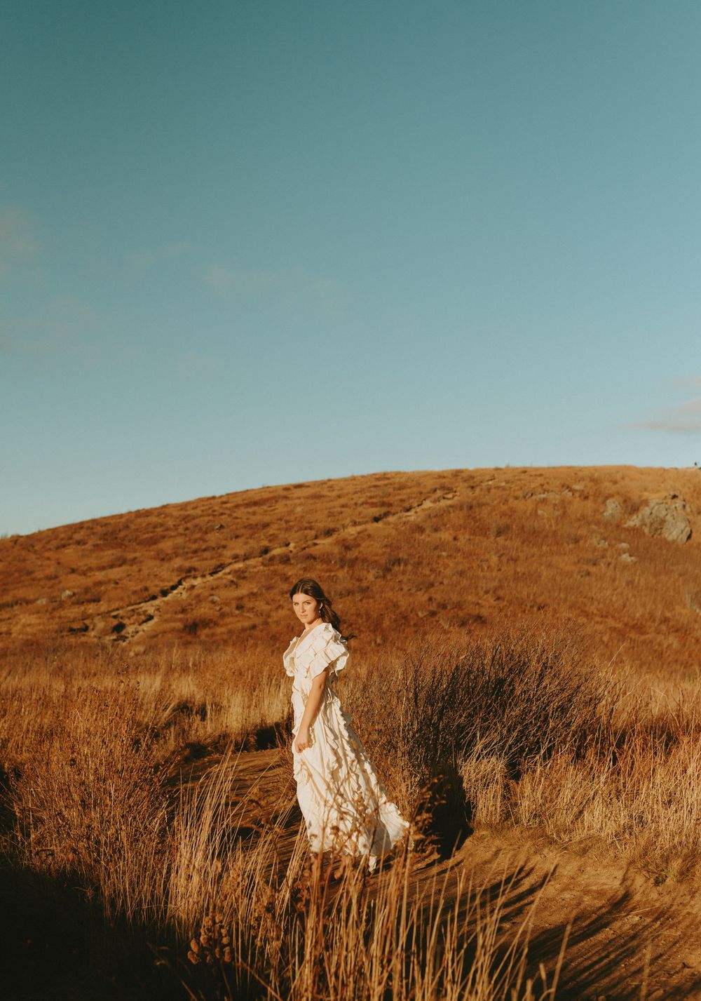 Person stands in tall golden grass against a blue sky on a hillside during golden hour.