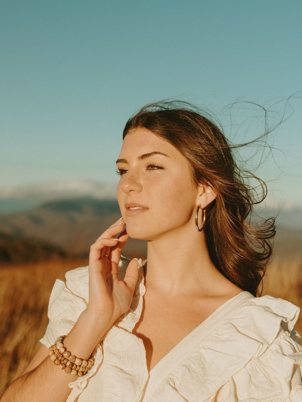 A person in a white blouse poses against a desert landscape with wind-swept hair at golden hour.