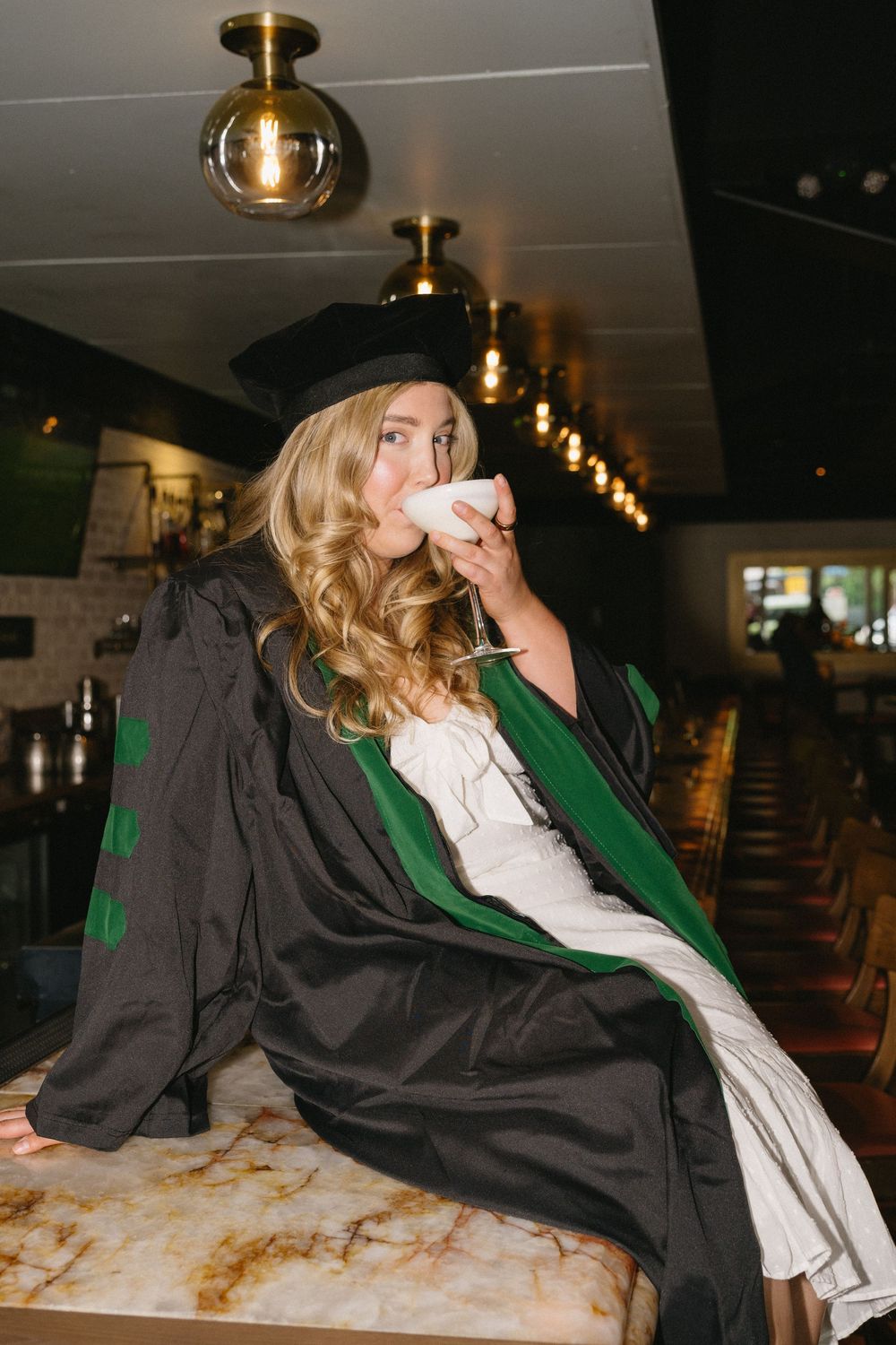 A person in a graduation gown and cap leans back dramatically in a series of artistic poses against a white surface.