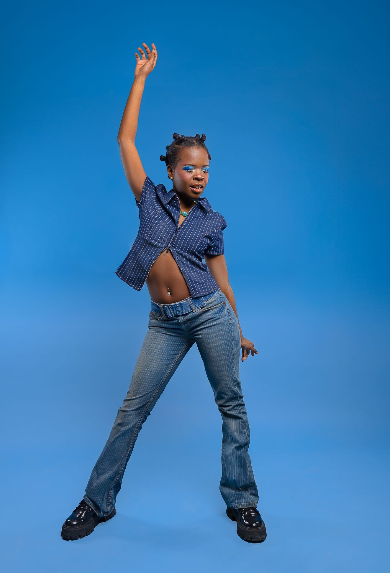 Photo of a girl in a full jean blue outfit, in front of a blue backdrop, mid dance, one arm up in the air, looking right