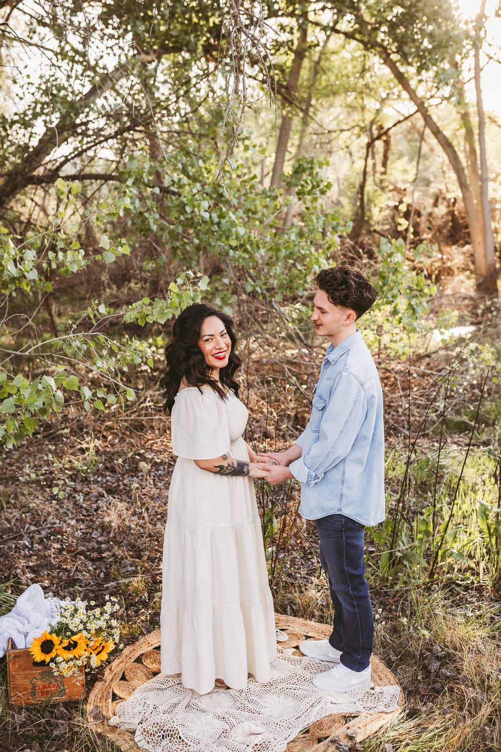 A couple shares a romantic moment in a sunlit wooded area with white dress and denim accents and yellow flowers.