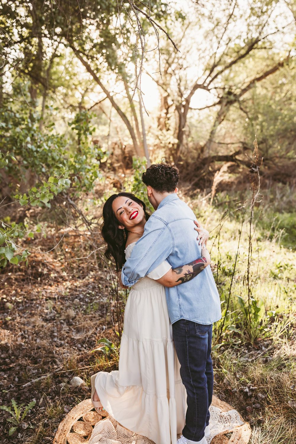 A romantic couple embraces and laughs together on a sunlit nature trail surrounded by trees and foliage.