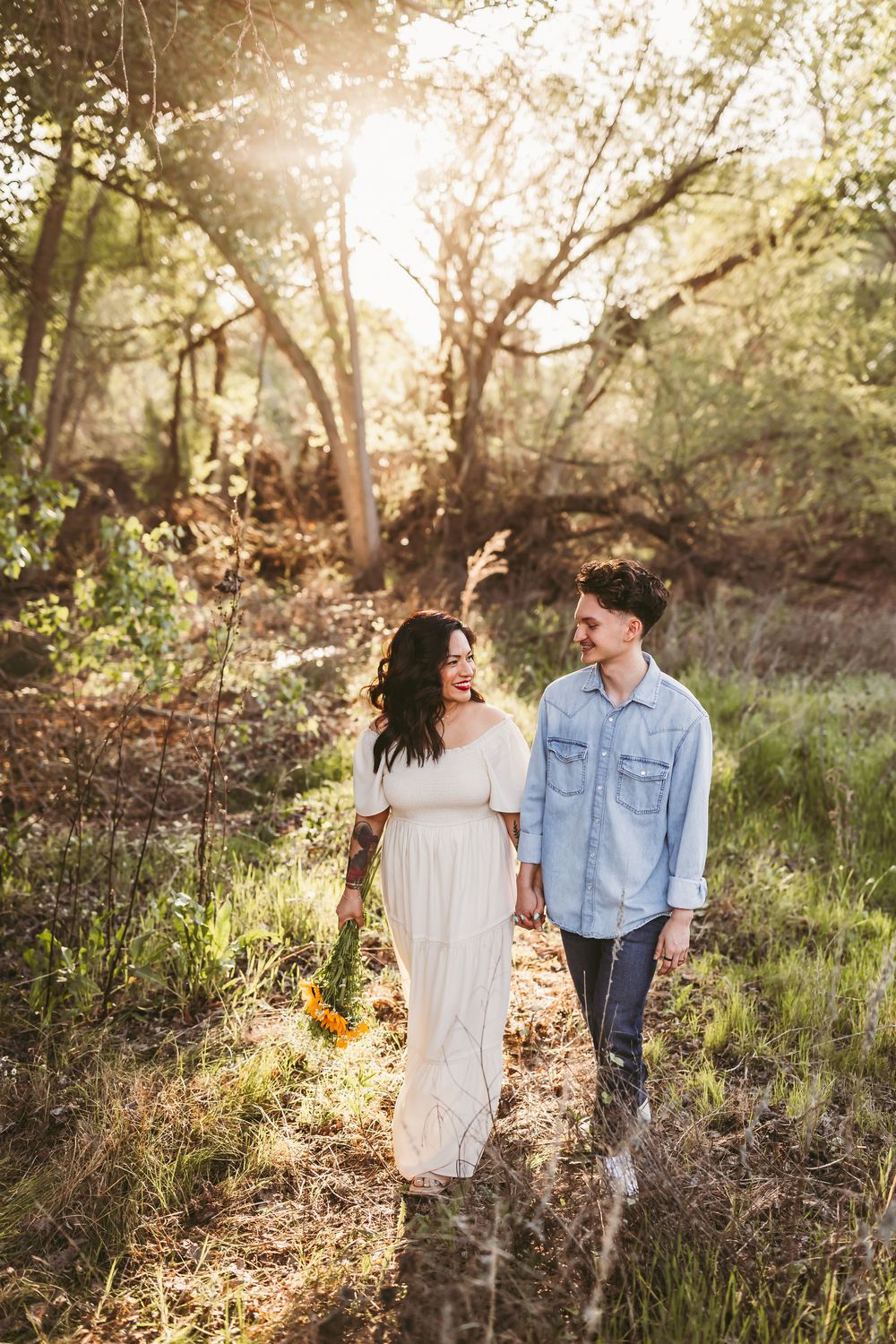 A couple walks hand in hand through a sunlit wooded area during golden hour, with the woman holding sunflowers.