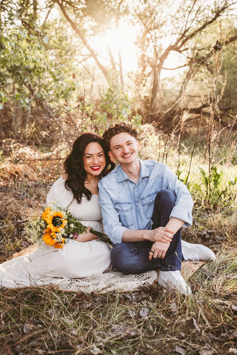 A couple relaxes on a blanket in a sunlit wooded area with sunflowers during an outdoor photo session.