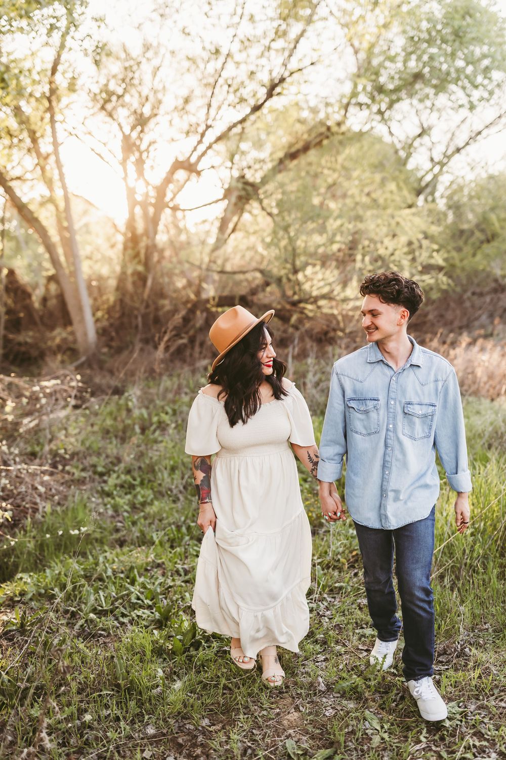 A couple takes a romantic stroll through a wooded area at golden hour wearing casual outfits.