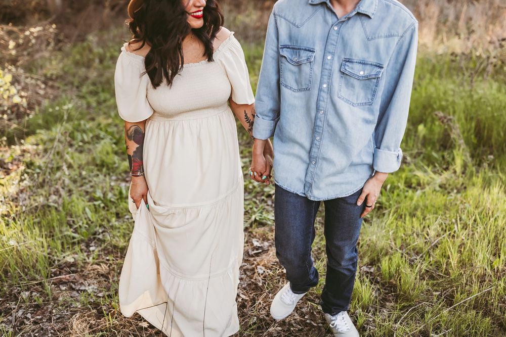 A couple stands together in a field wearing casual attire - a flowing white dress and denim shirt with jeans.