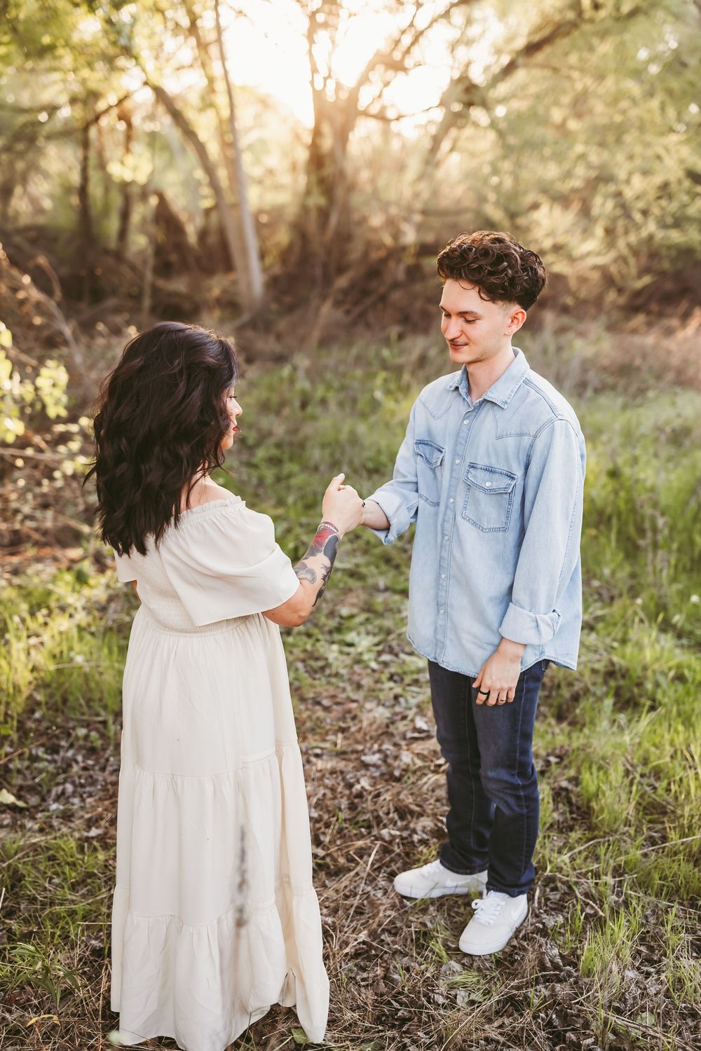A couple in white dress and denim embraces while walking through a wooded area during golden hour.