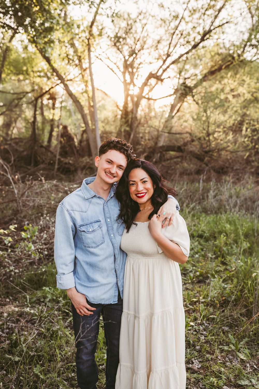 A couple shares a romantic moment in a sunlit wooded area wearing coordinating blue and white outfits.