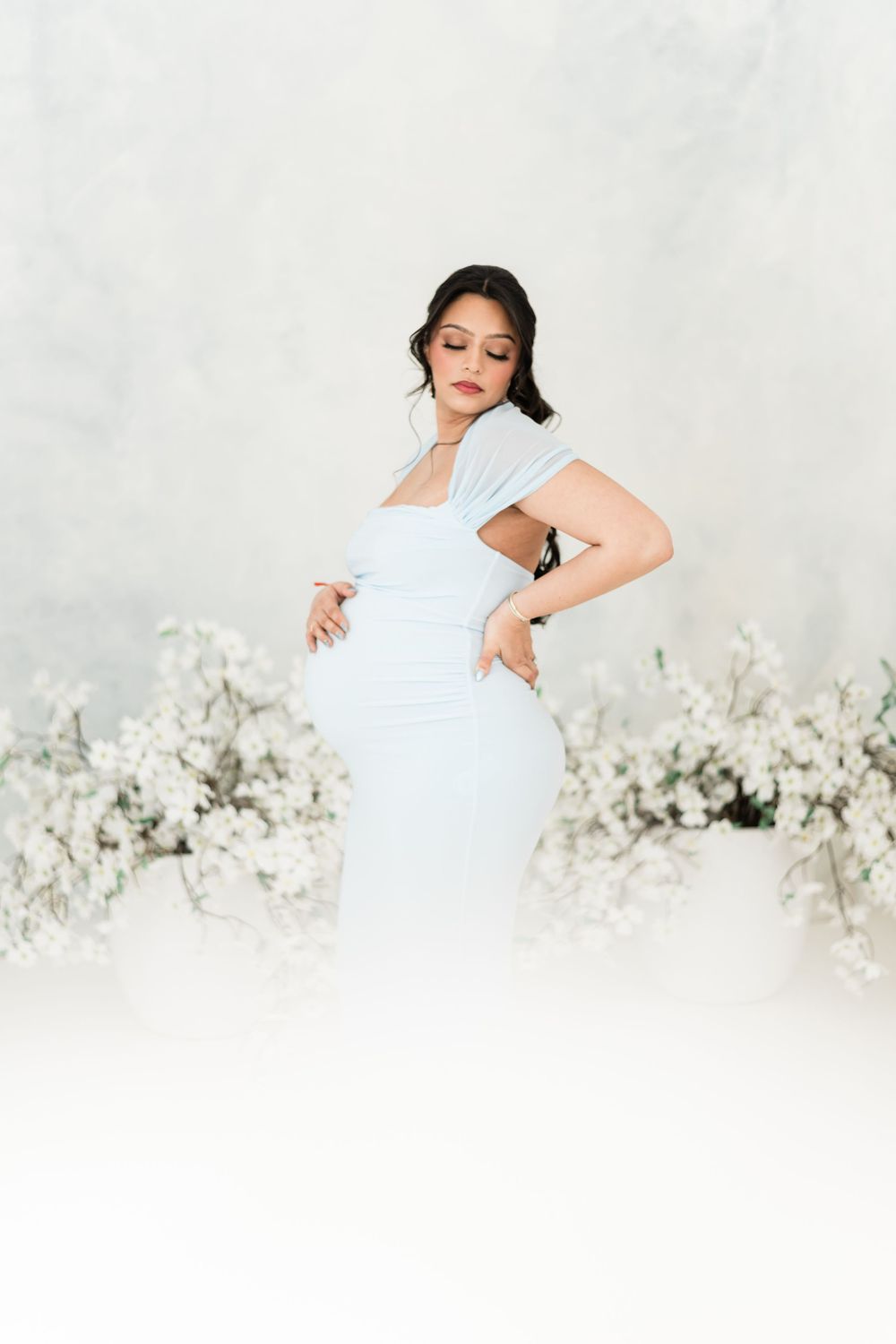 Pregnant woman in light blue dress surrounded by white flowers.