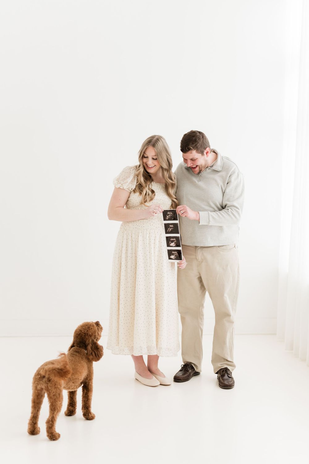Expecting couple holding ultrasound photos with their brown poodle in a bright white studio.