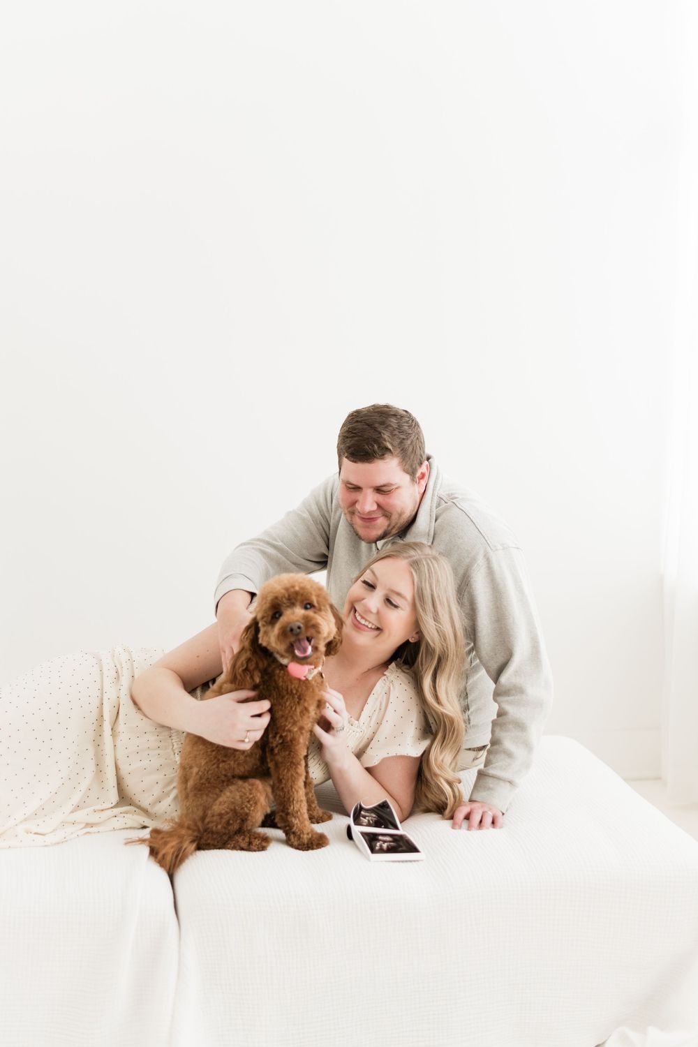 Smiling couple with brown poodle and ultrasound photos lying on a white couch.
