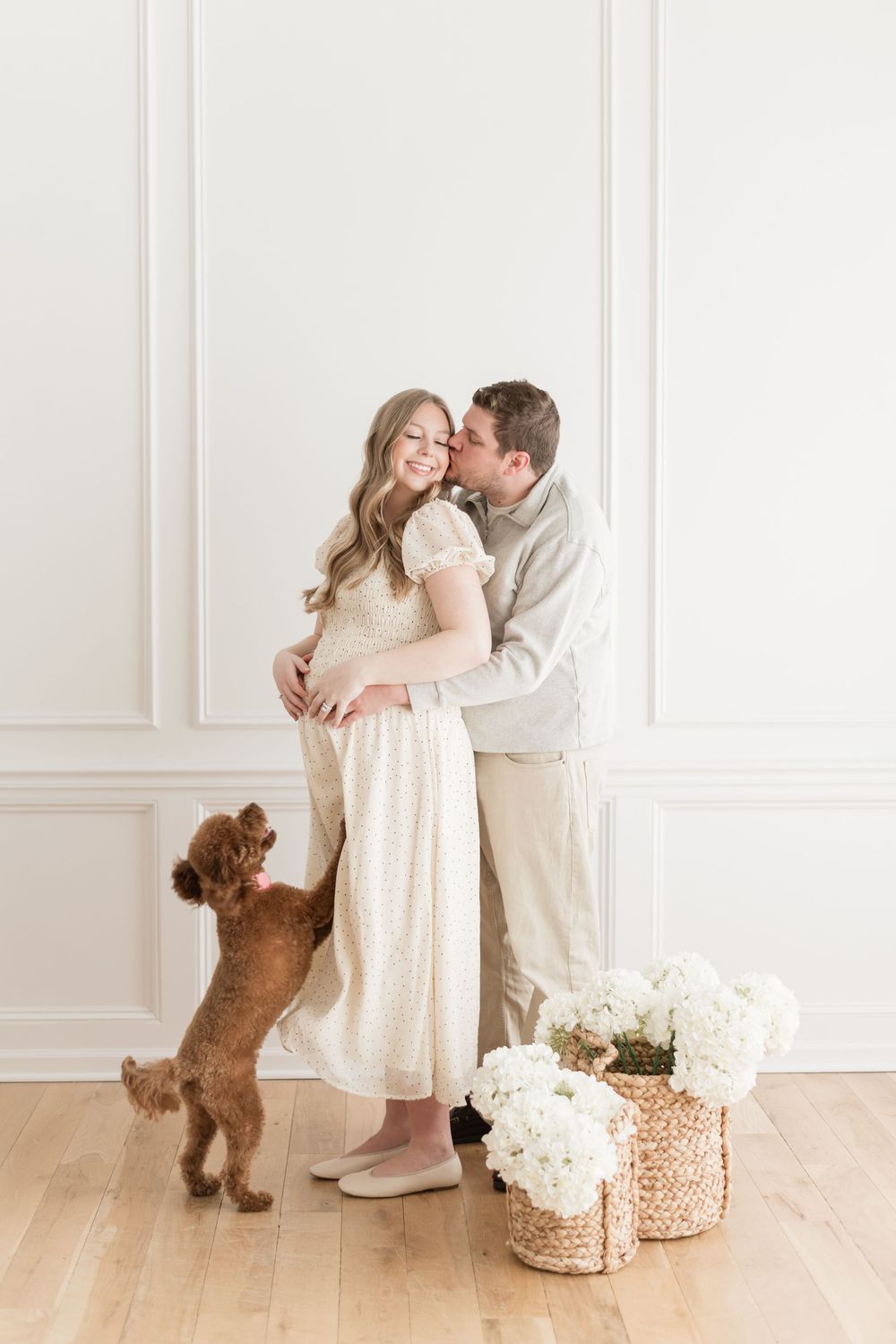 Couple embracing while brown poodle jumps up beside them near white hydrangea baskets.