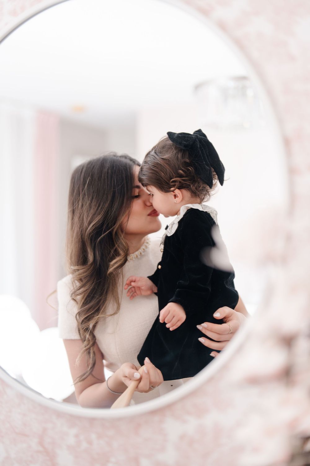 Mother kissing her baby girl in a black dress, reflected in a round mirror.
