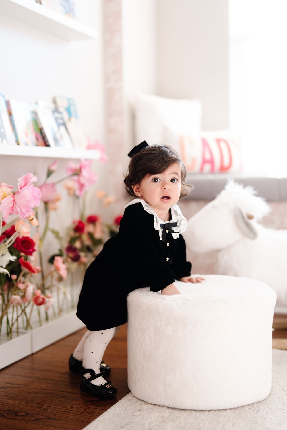 Adorable baby girl in black velvet dress standing by a white ottoman in a bright room.