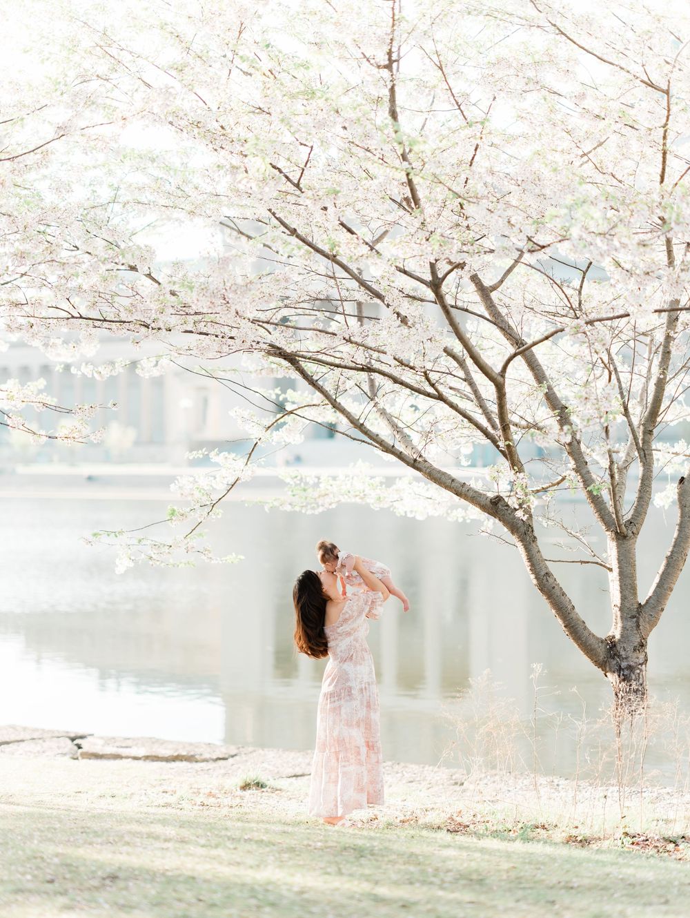 Mother lifting baby under blooming cherry blossom tree beside a tranquil waterway.