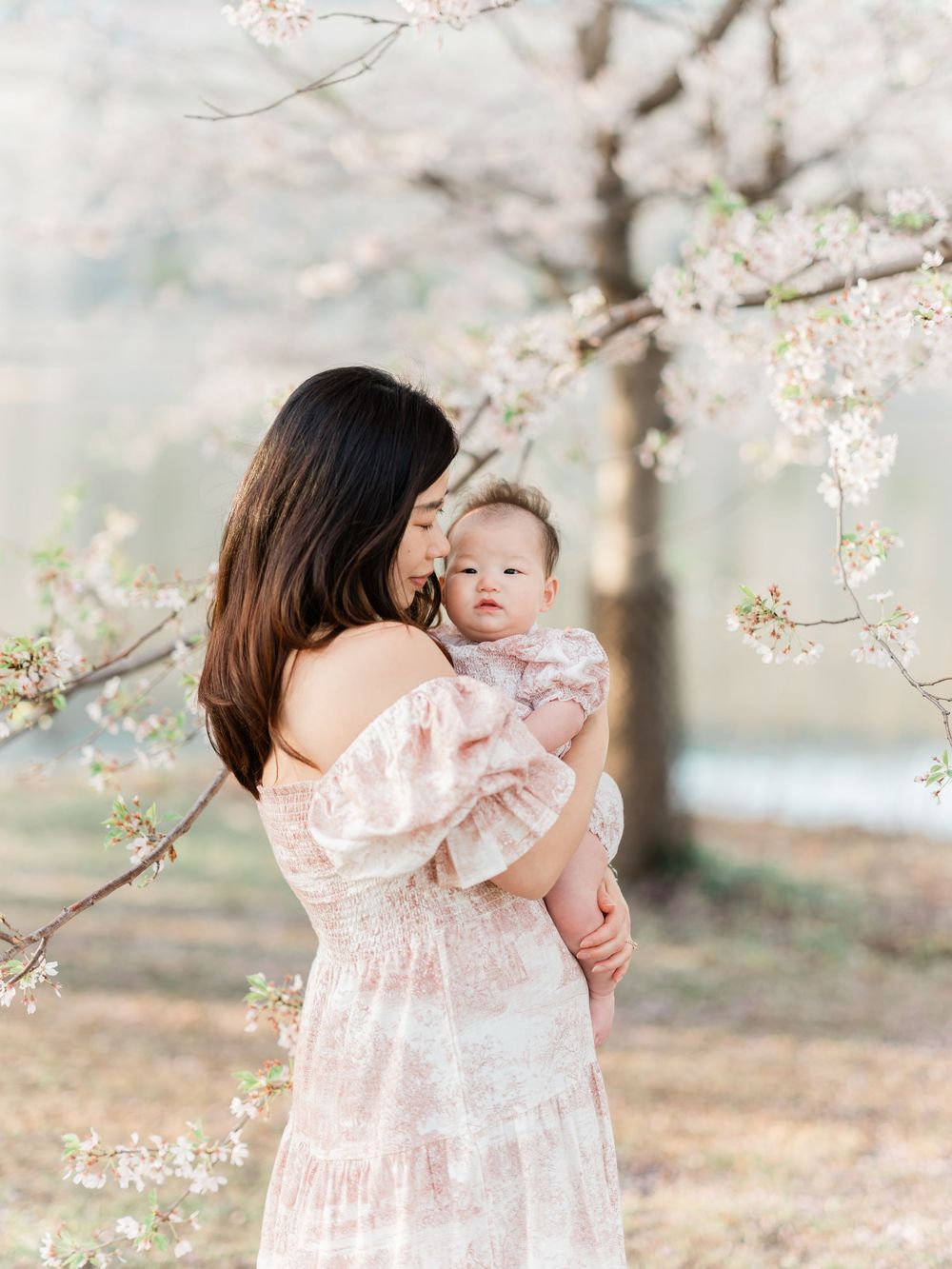 Mother in floral dress kissing baby amid cherry blossoms.