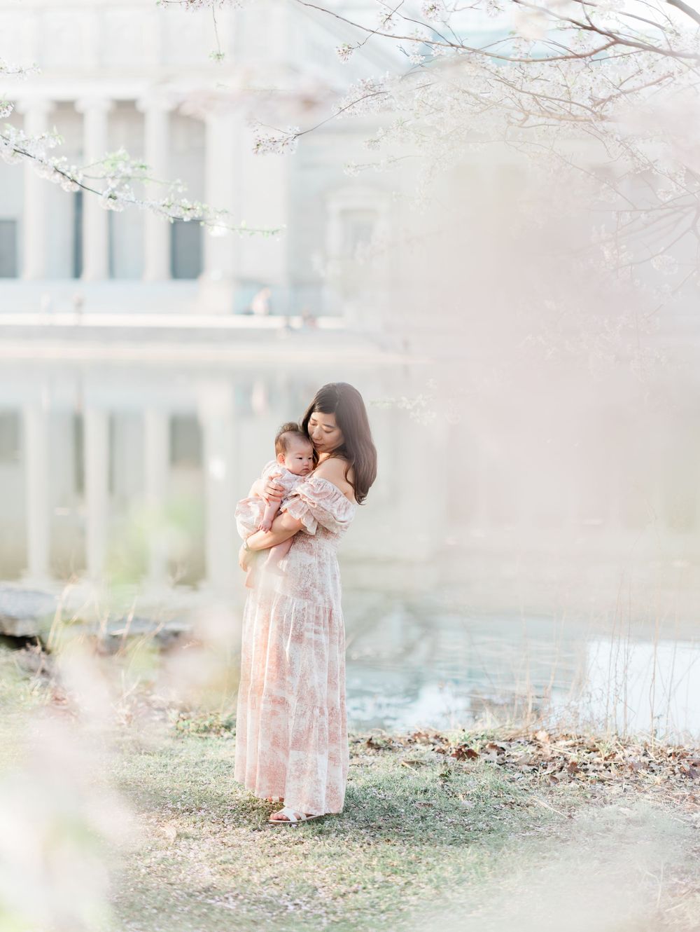 Mother holding baby near cherry blossom trees by a pond.