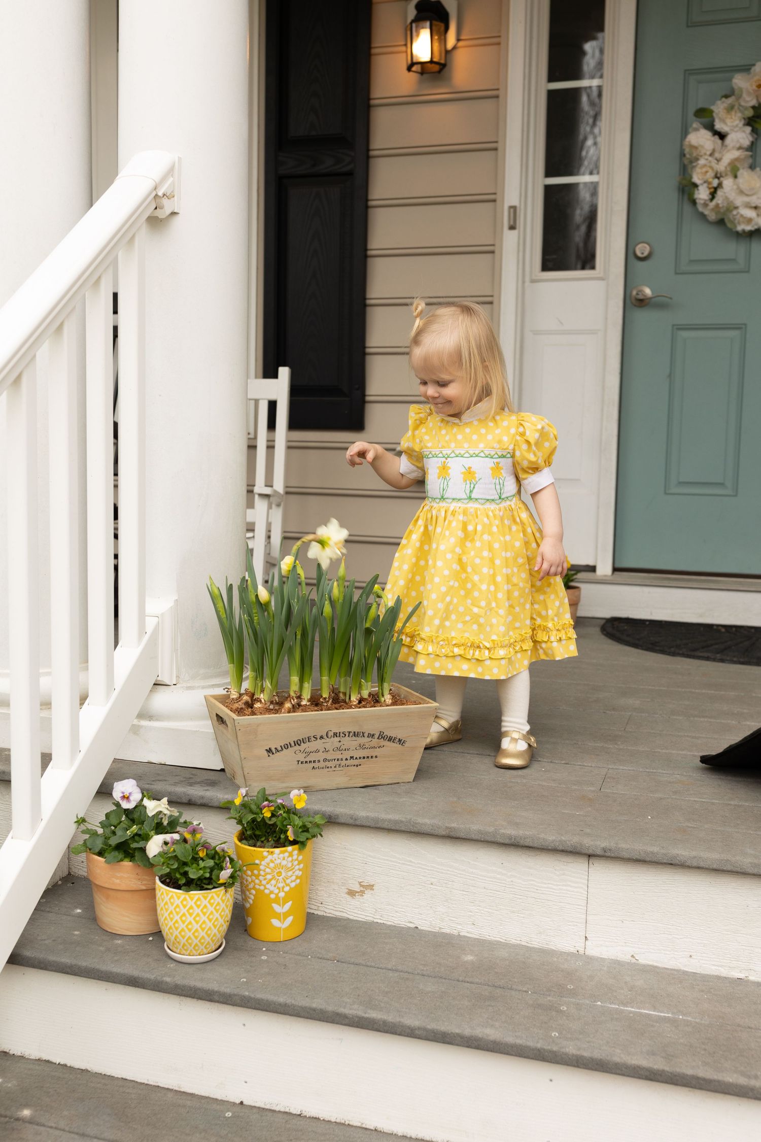 Little girl in a daffodil dress reaches toward a crate of daffodils on her front porch, by Charlottesville family and baby photographer.