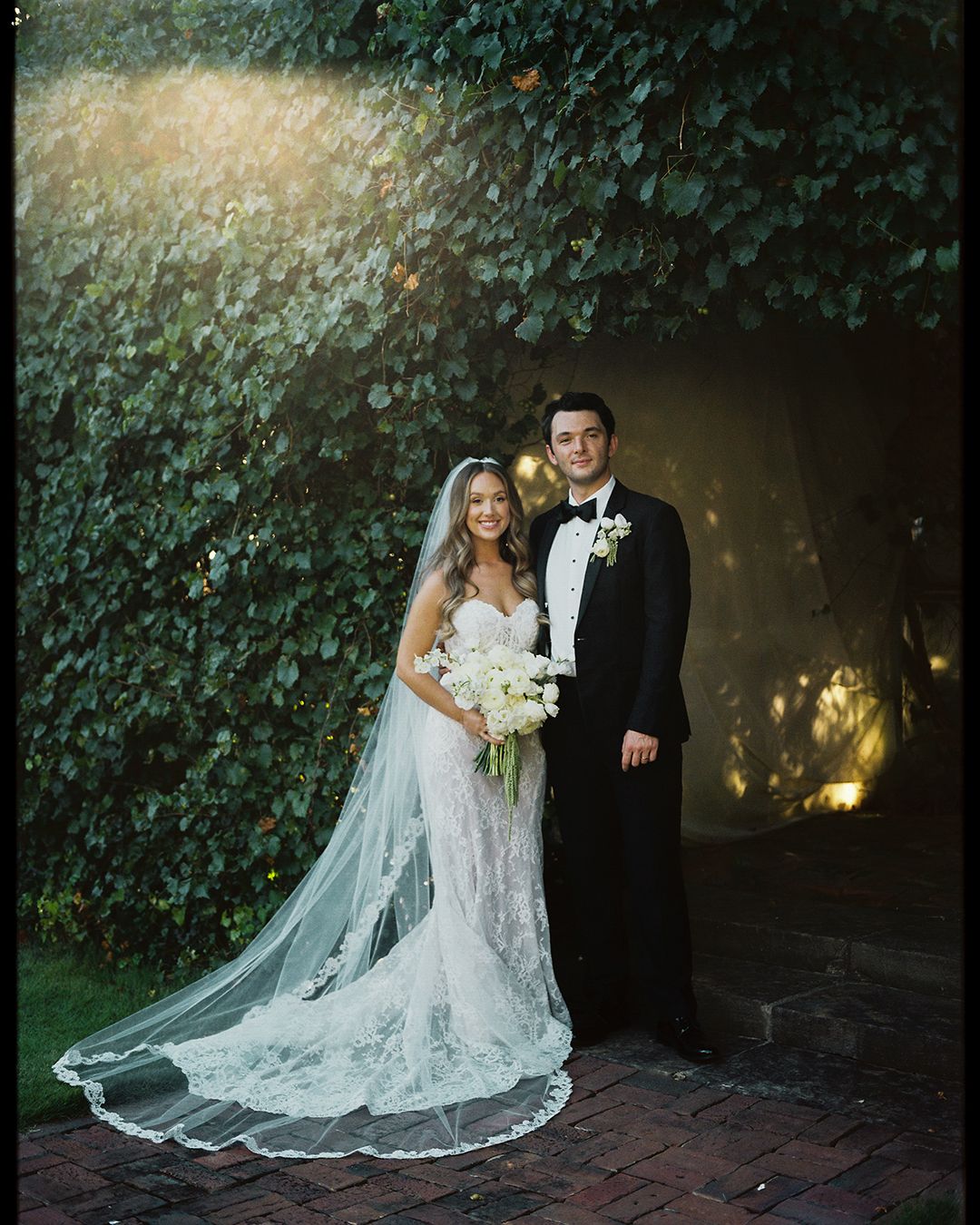 Bride and groom pose elegantly in front of an ivy-covered wall during their romantic evening wedding photoshoot.