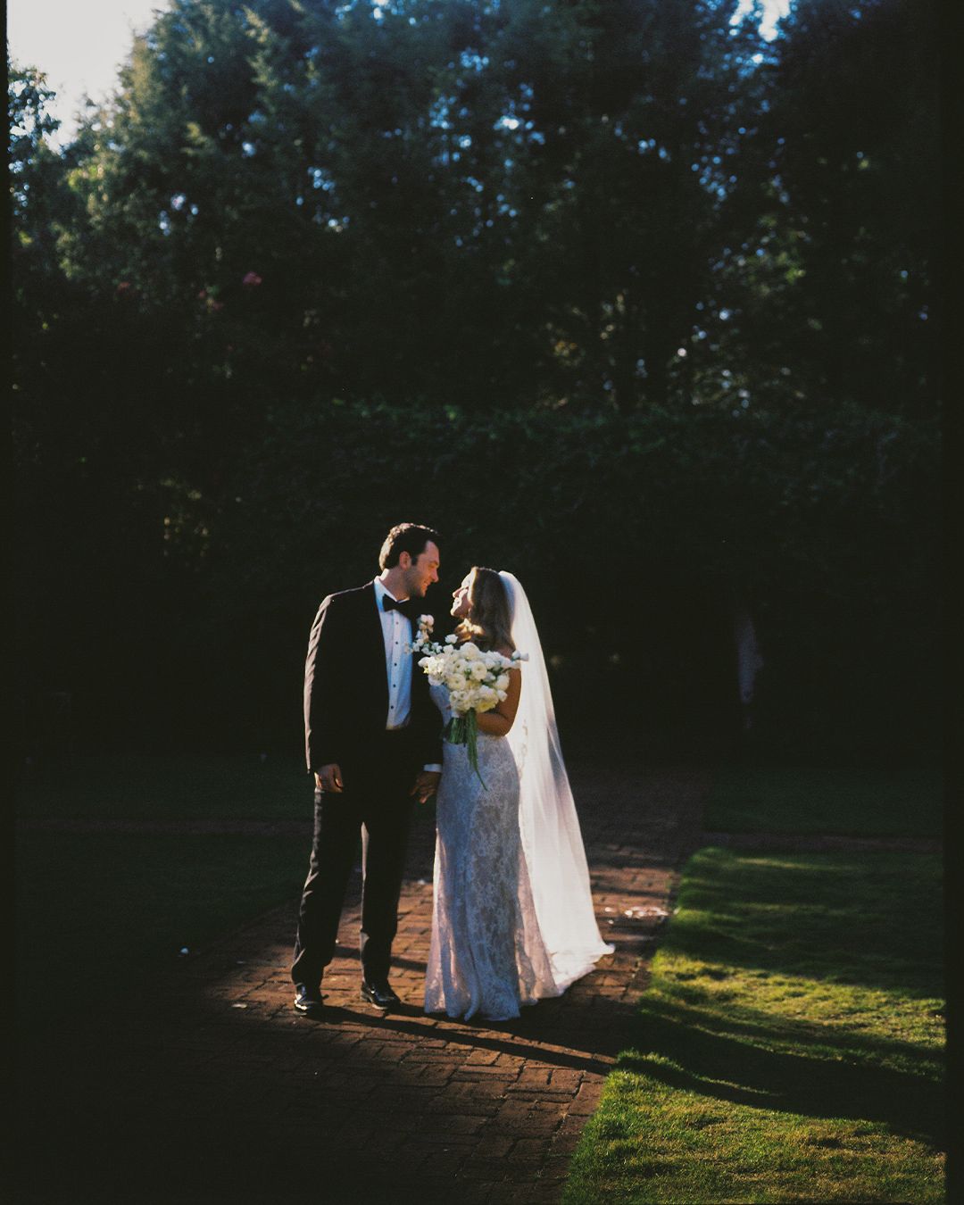 A couple in wedding attire walks together in a sunlit forest clearing during golden hour.