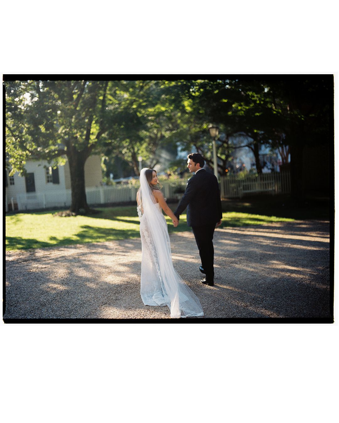 A couple in wedding attire walking together on a tree-lined path during a romantic outdoor photo session.