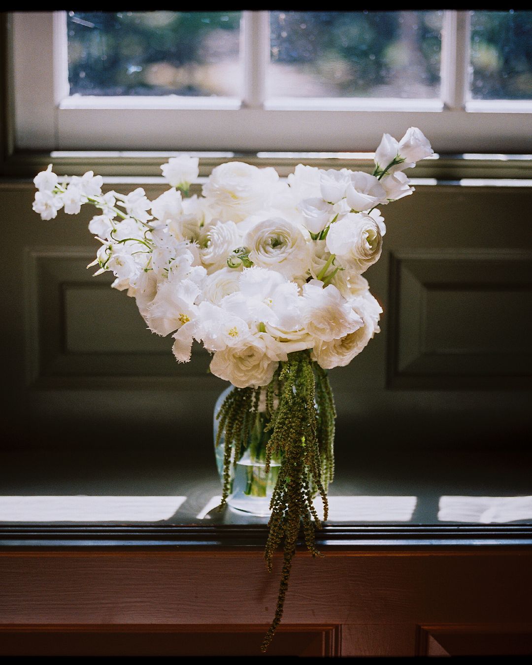 A glass vase filled with white ranunculus and roses sits on a windowsill, creating an elegant floral arrangement.