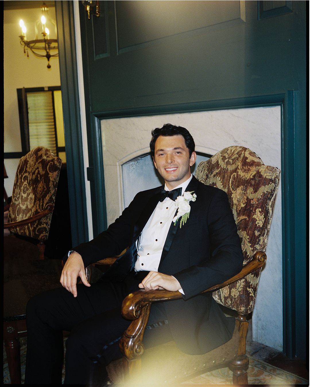 A person in a black tuxedo poses on a vintage floral patterned sofa in a dimly lit room.