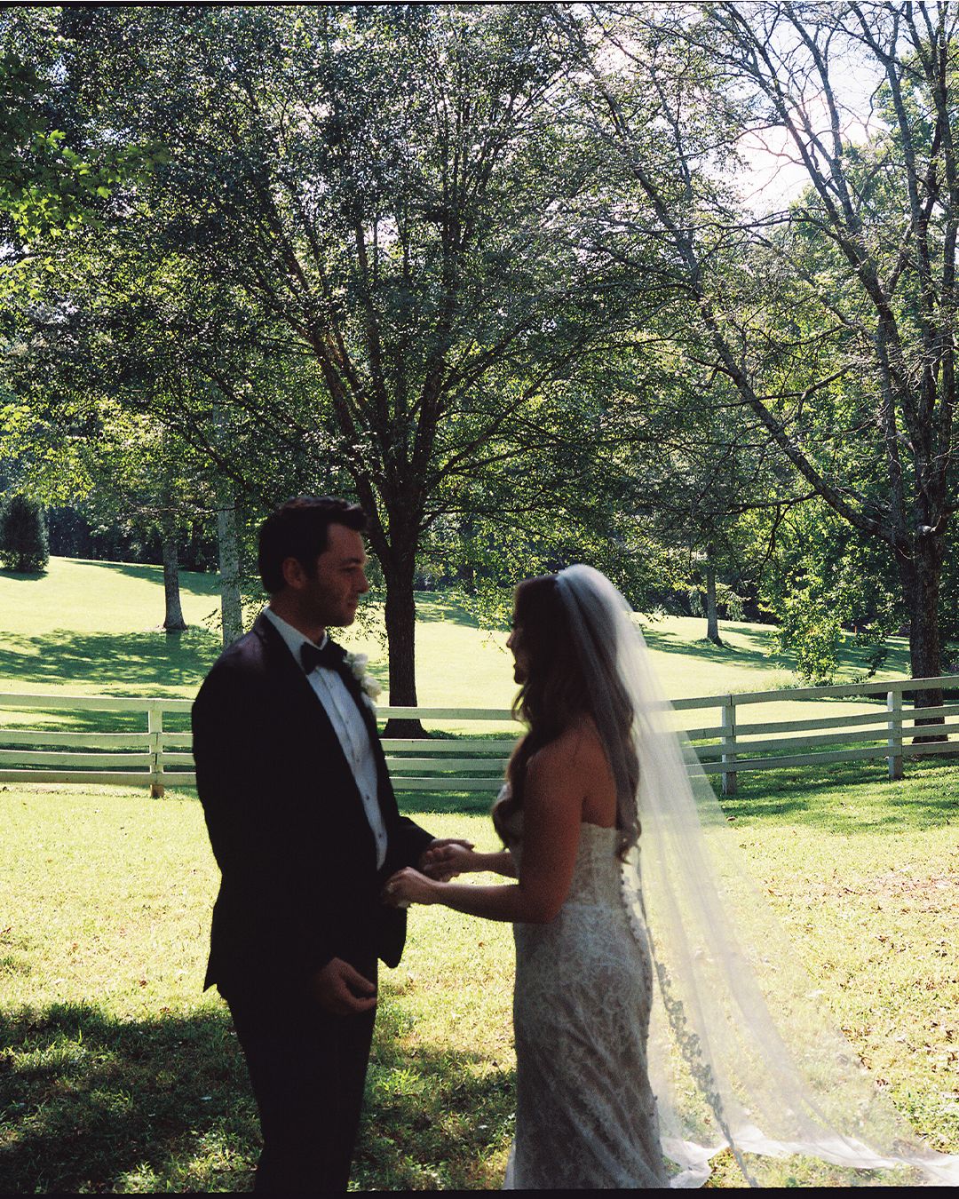 A couple shares an intimate moment during their outdoor wedding ceremony surrounded by lush green trees in a park setting.