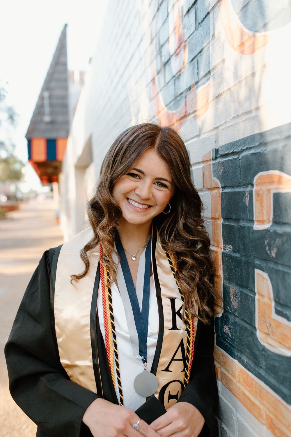 A graduate in cap and gown with honor cords poses against a brick wall.