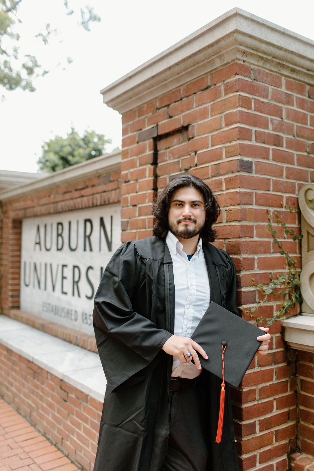 A graduate in black academic regalia holds diploma in front of Auburn University sign.