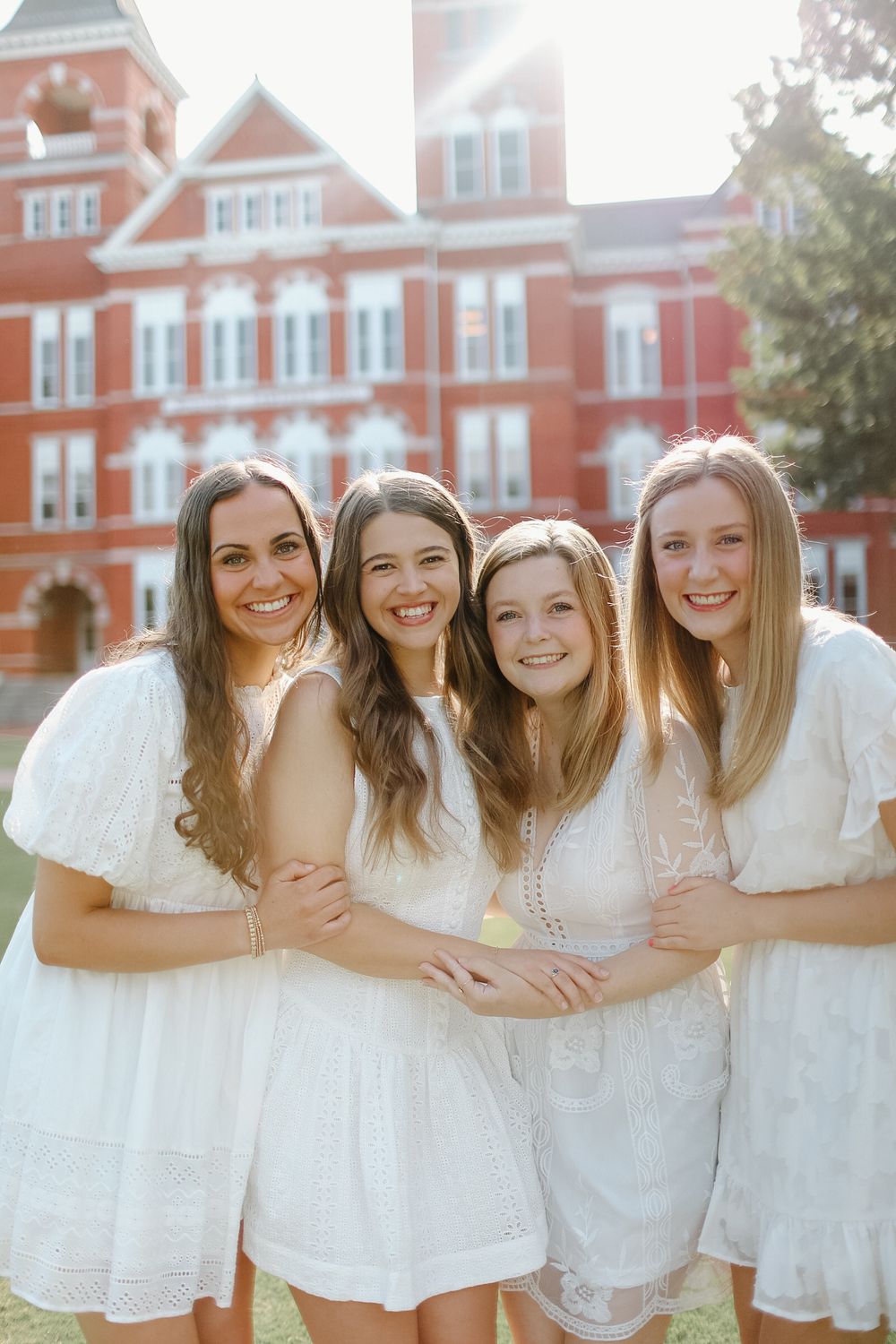 A group of friends in white dresses pose together smiling in front of a red brick university building.