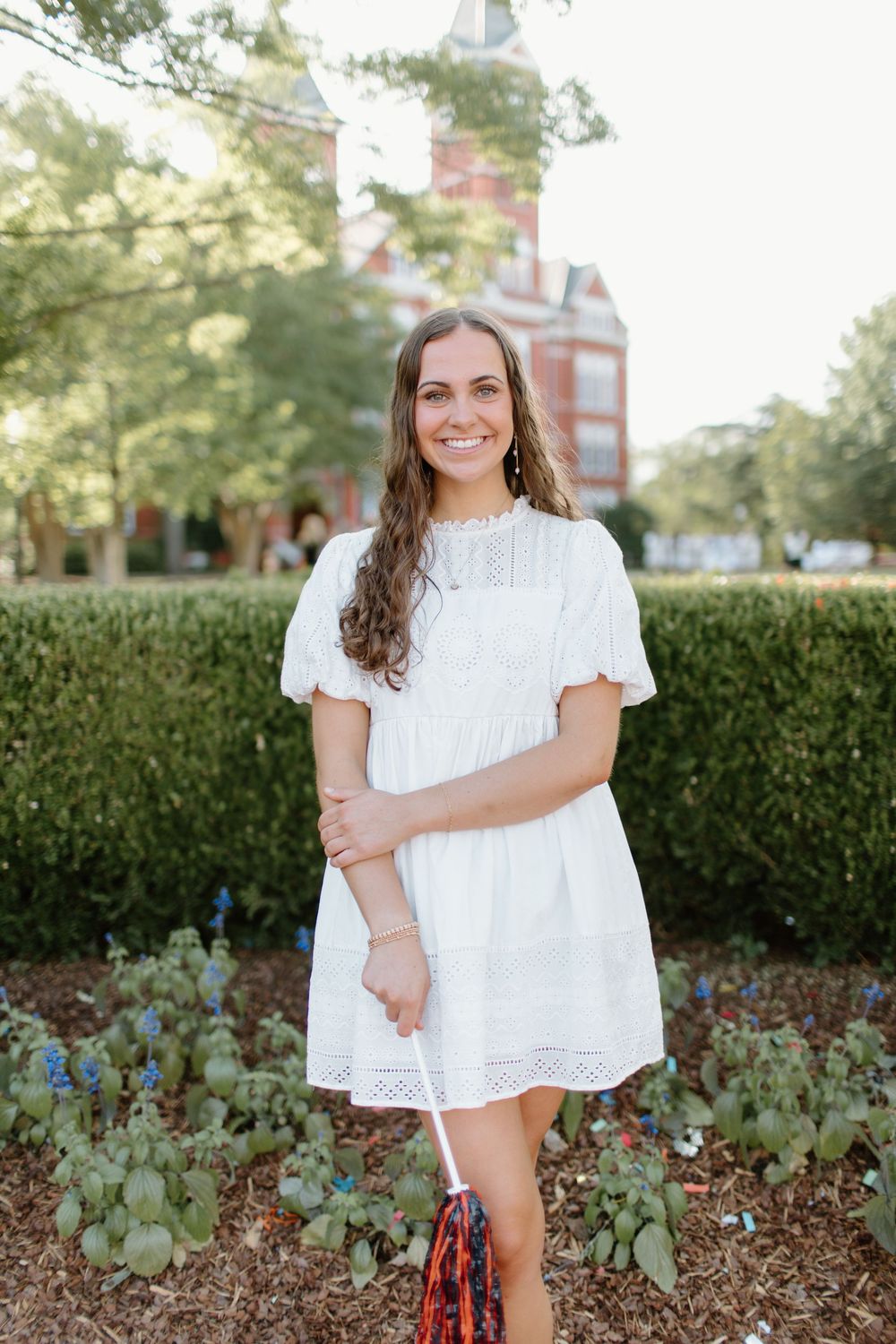 A person in a white summer dress poses outdoors near a red brick building and garden.