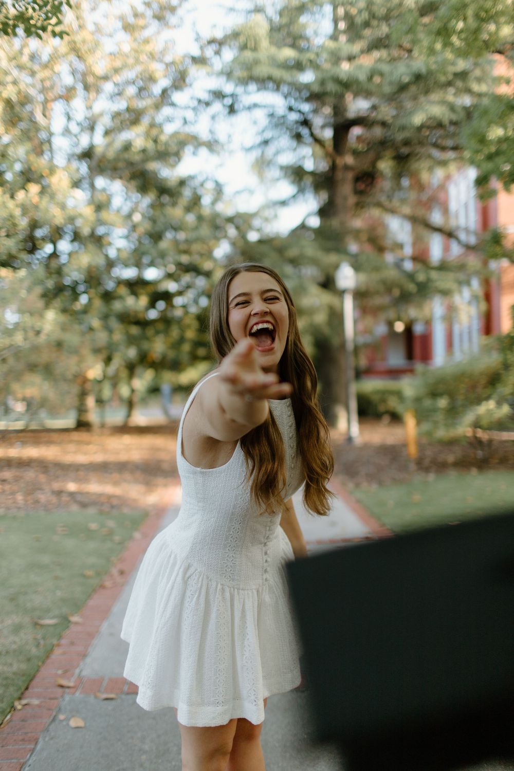 A person in a white dress laughs joyfully while posing on a brick pathway.