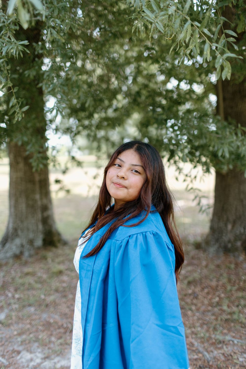 A person in blue graduation attire poses outdoors among trees.