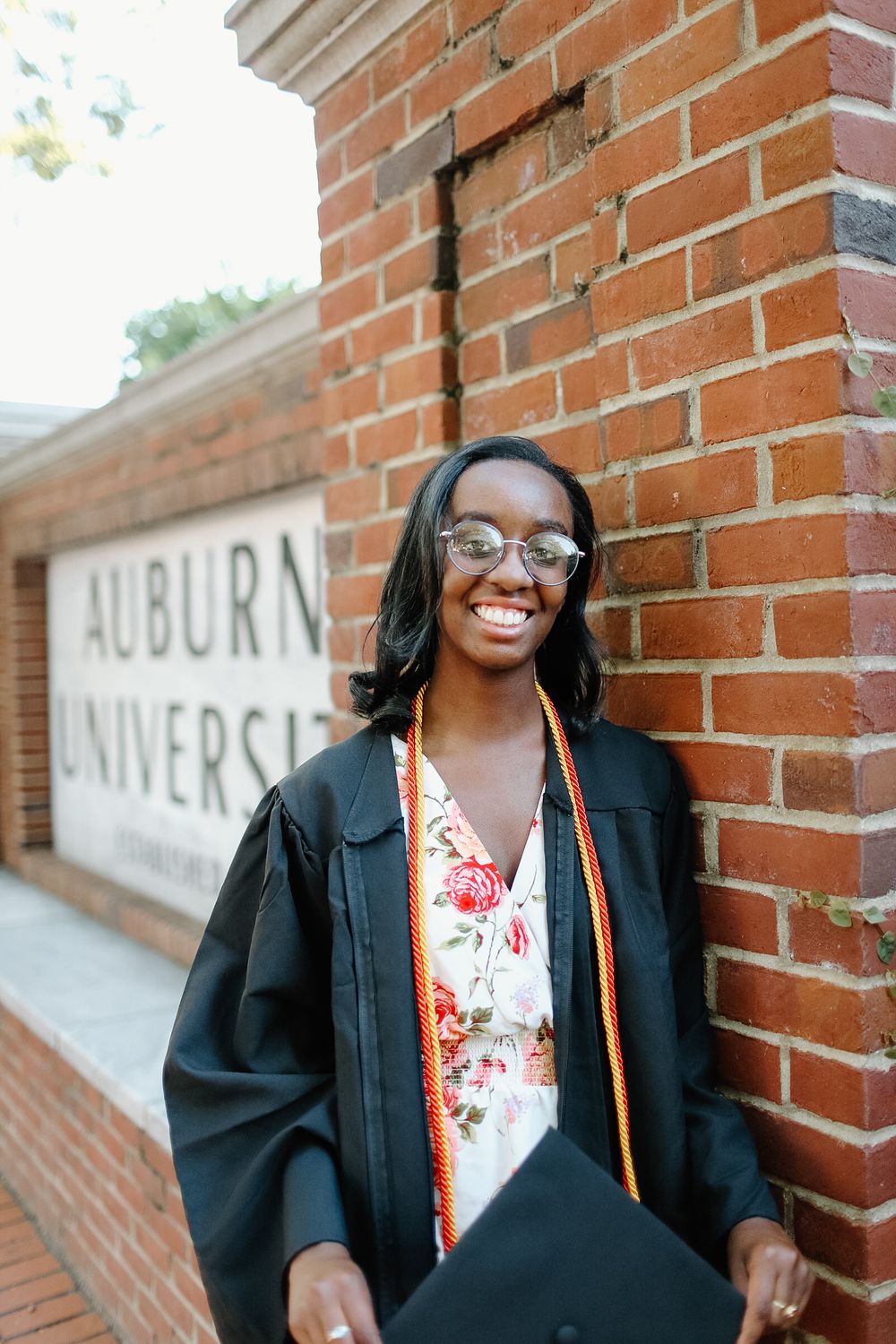 A graduate in academic regalia smiles brightly in front of an Auburn University sign.