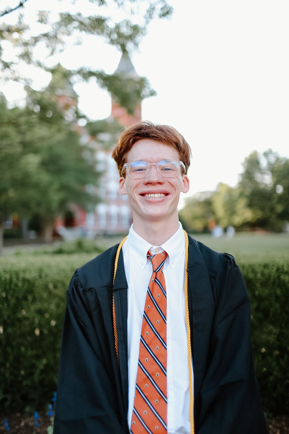 A graduate in cap and gown with striped tie poses outdoors on campus.