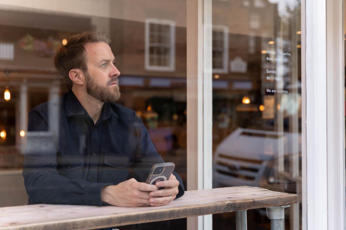 Personal branding photograph taken through a window of a life coach holding his mobile phone, looking out into the distance